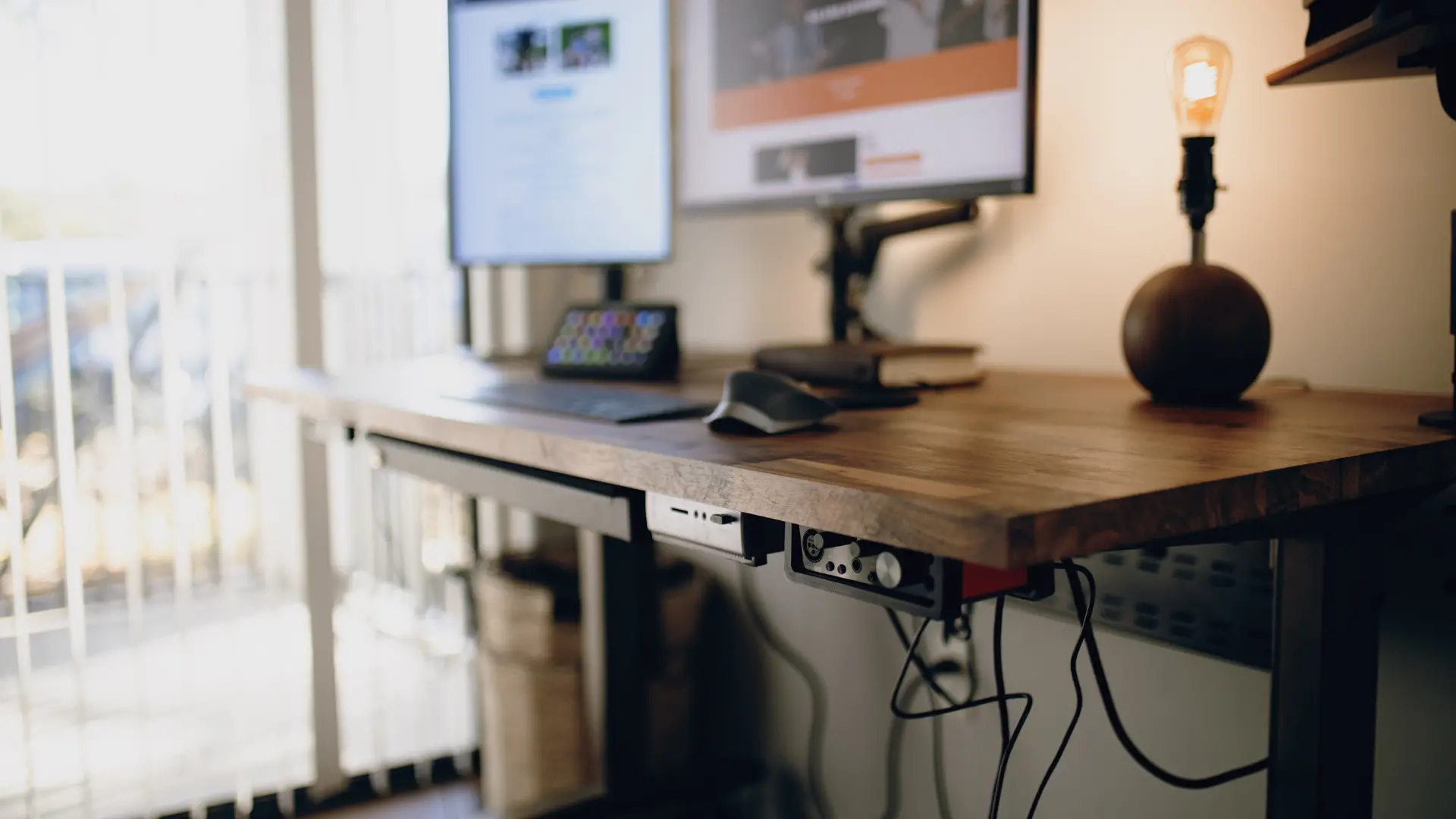 A timber-topped adjustable desk with a monitor, keyboard and desk lamp beside a bright window.