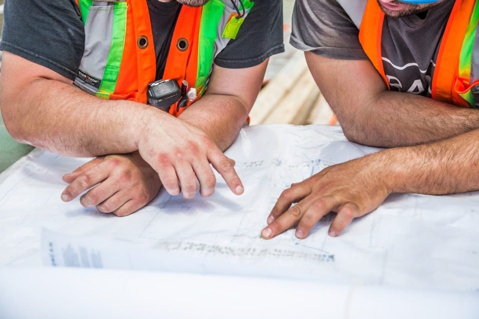 Two construction workers in safety vests reviewing printed drawings