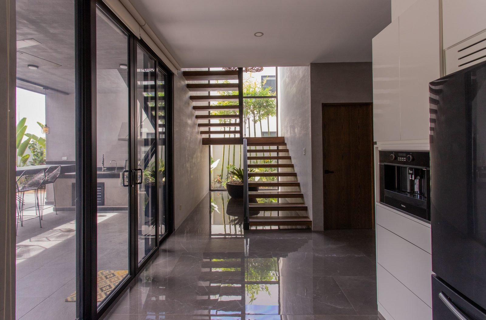 Model home interior with a floating timber staircase, glass sliding doors, polished floors and built-in kitchen cabinetry.