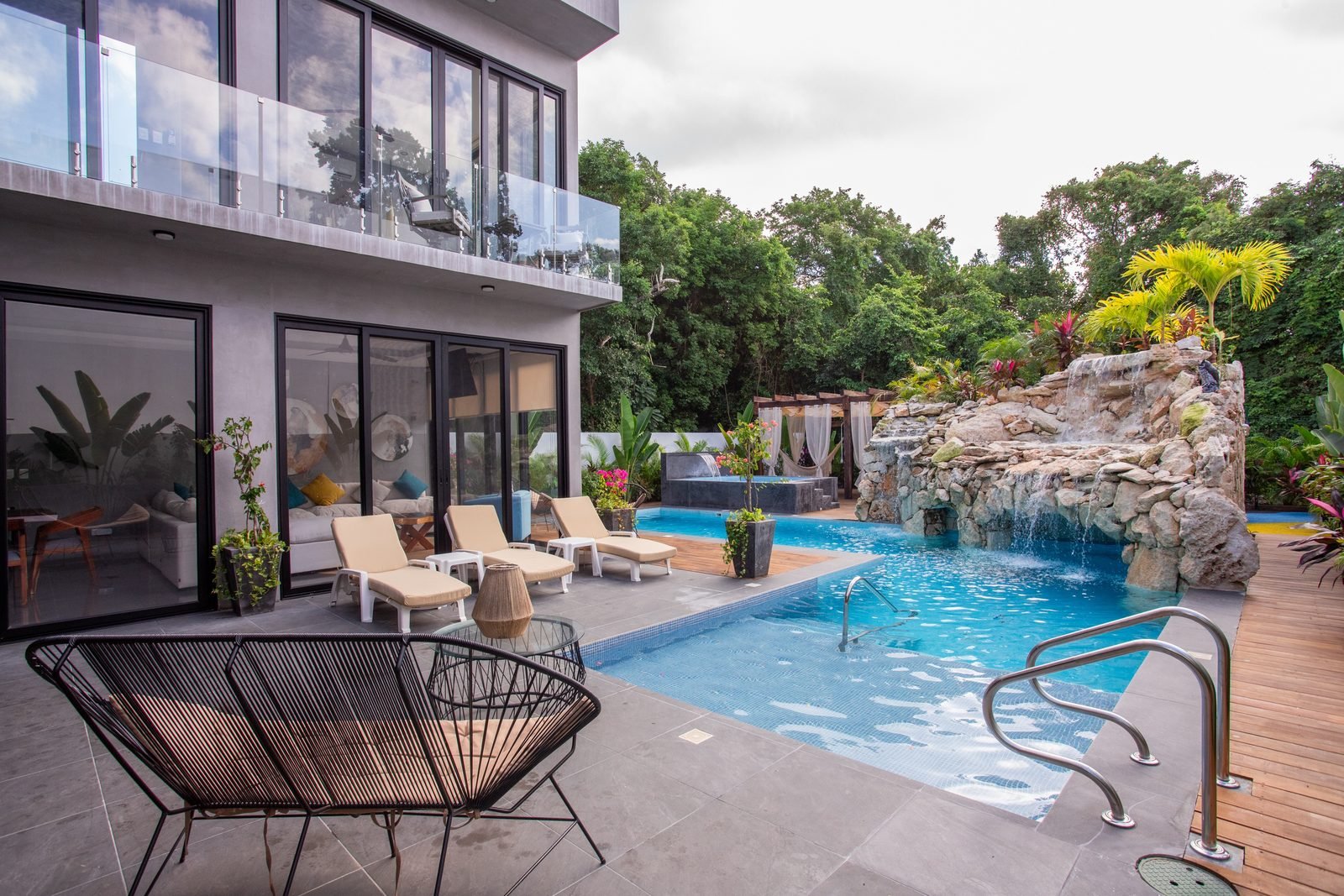 Model home terrace with a swimming pool, rock grotto waterfall, sun loungers and glass balcony railings.